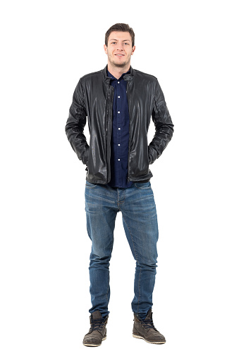Relaxed man in jeans and leather jacket smiling at camera with hands in pockets. Full body length portrait isolated over white studio background.