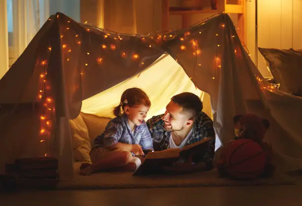 Photo of happy family father and child daughter reading a book  in  tent at home