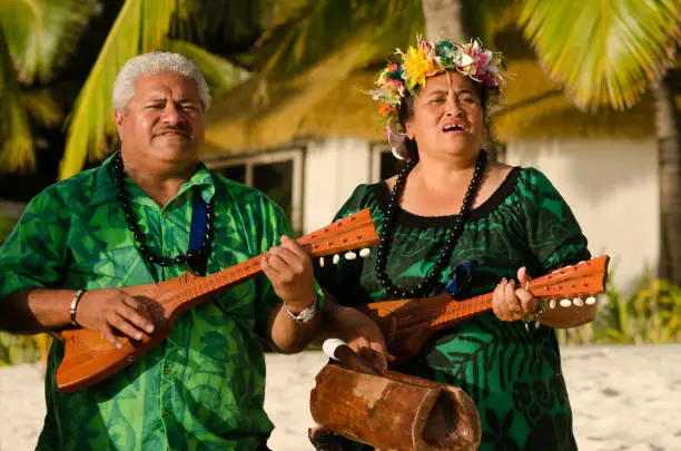 Portrait of two mature Polynesian Pacific islanders couple sing and plays Tahitian Music with Ukulele guitars on tropical beach with palm trees in the background.