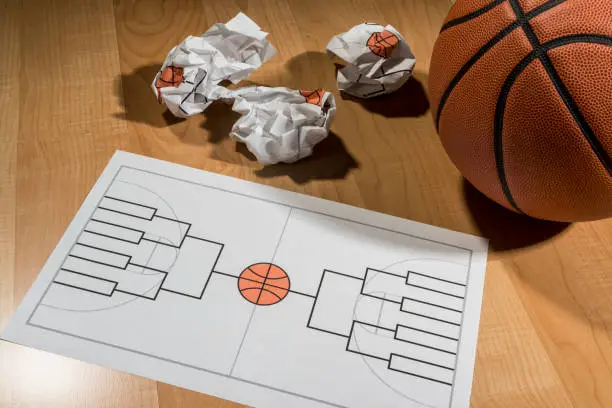 Attempting to fill out a college basketball tournament bracket on a legal size piece of paper with several crumpled balls of paper with failed attempts, sitting on a basketball court with a basketball sitting next to it. This bracket is for a 16 team single elimination tournament, winner take all,  which typically happens in March every year in the US between college basketball teams and many people gamble on the series.