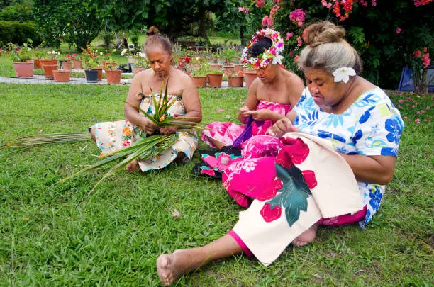 Portrait of Polynesian Pacific Island Tahitian mature females sewing Tivaivai and weaving a hat knees out of palm leaves outside their home in Aitutaki lagoon Cook Islands.