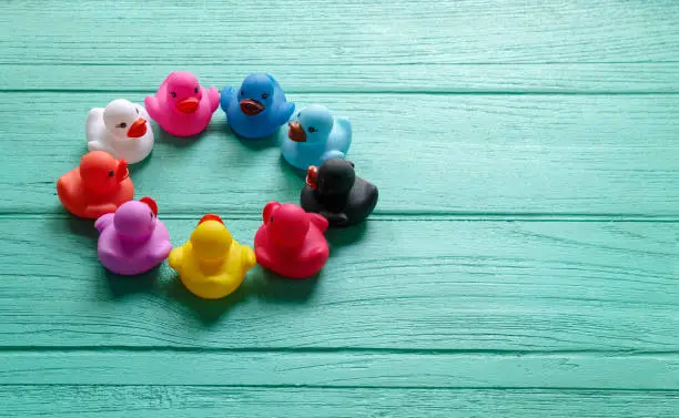 A group of rubber ducks of different colors sit in a circle opposite each other as if discussing and talking about their differences but being together peacefully on an old wooden turquoise colored grained table. Concept image representing discussion, negotiation, peace, living together, race, gender, ethnicity, living in harmony etc.