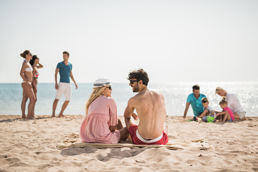 Back view of young smiling couple talking to each other on the beach, while other people are in the background.