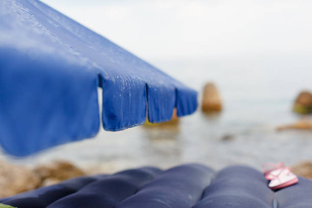 Rain raindrops flow down the beach umbrella, which stands on the beach overlooking the sea and rocks. Near the inflatable mattress, on which lie pink children's flip flops. Summer, the sea.