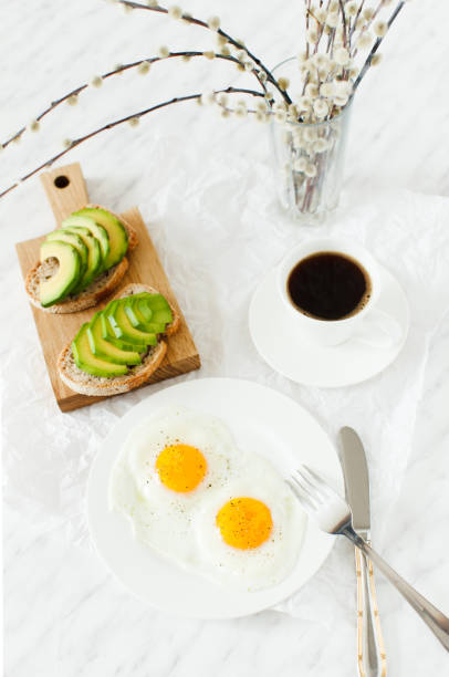 istock Morning breakfast, table setting. On a white plate are two fried eggs, on a wooden board sliced avocado, a cup of coffee and a vase with a plant. White background, daylight, vertical image. 655749326