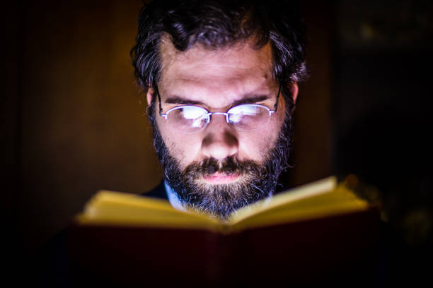 Handsome, middle age, bearded man reading book in his home under the lamp light. Close up. He is focused. Shallow DOF, focus on eyes
