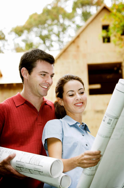 istock Young couple looking over construction blueprints with their home under construction  in the background. 655810302