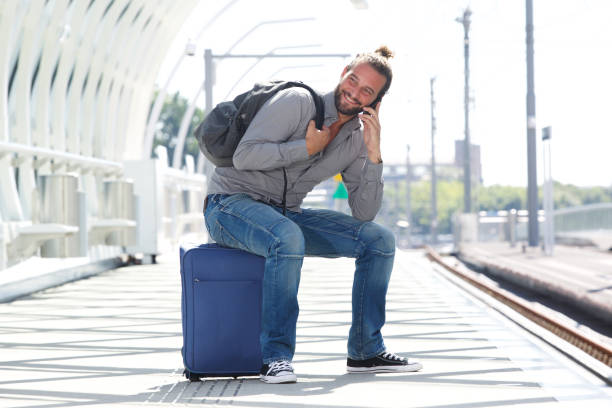 istock Smiling man sitting on suitcase and talking on cell phone 655812198