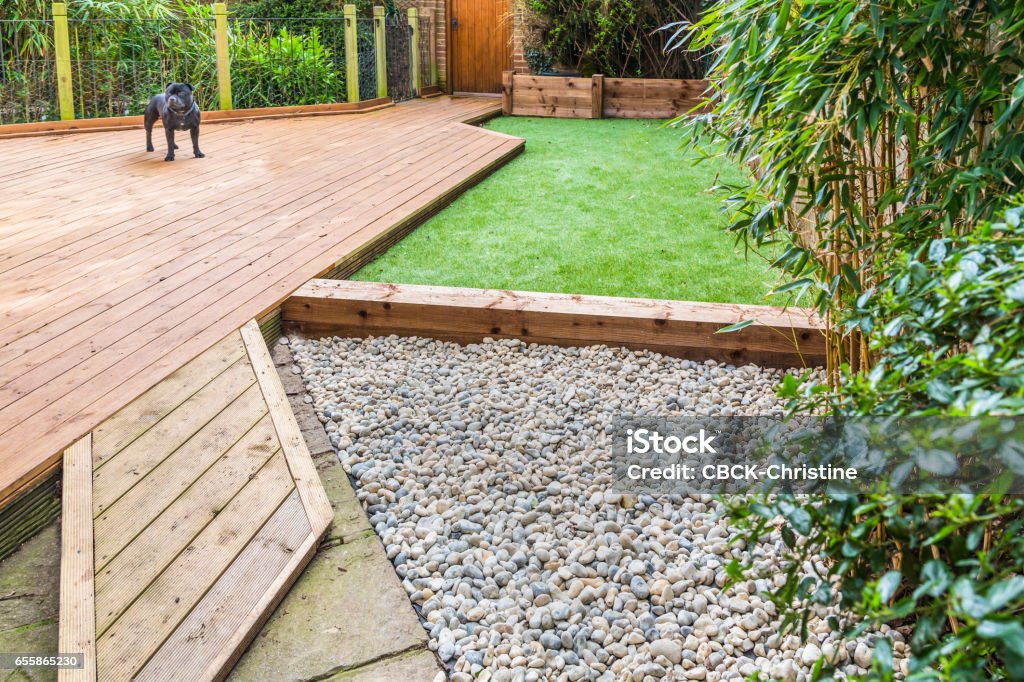 A section of a residntial garden, yard with wooden decking and artificial grass A section of a residntial garden, yard with wooden decking, patio over a fish pond, a section of artificial grass and an area of stone pebble. There is a bamboo plant and a dog in the garden. Artificial Stock Photo