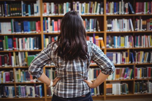 Rear view of female student looking at books in the shelf in the library