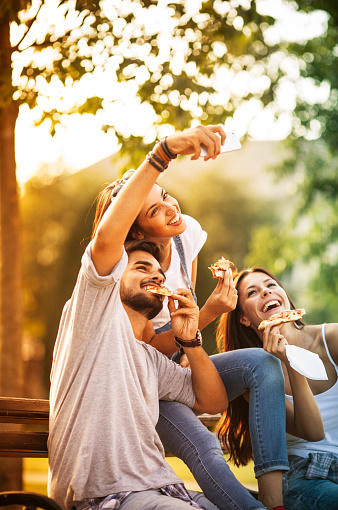 Group of young friends sitting on park bench and eating pizza.Taking selfie.