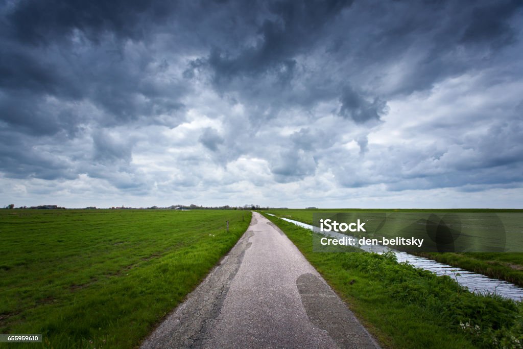 Road with dramatic blue cloudy sky and green grass in cold day in spring. Fantastic landscape in Netherlands. Nature and travel background. Rural road with green fields Dramatic Sky Stock Photo