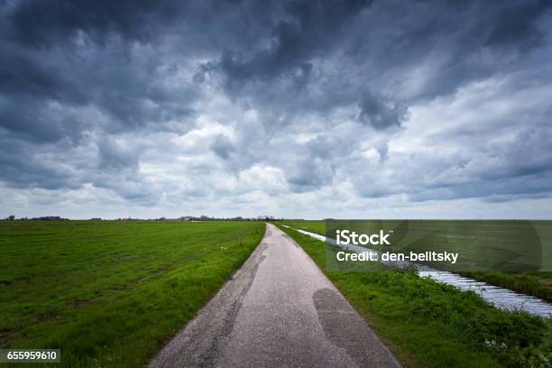Road With Dramatic Blue Cloudy Sky And Green Grass In Cold Day In Spring Fantastic Landscape In Netherlands Nature And Travel Background Rural Road With Green Fields Stock Photo - Download Image Now