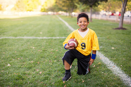 A young boys dressed in a flag football uniform stands posed for a portrait ready for a great game. He is smiling and looking at the camera while standing on a football field in Utah, USA.