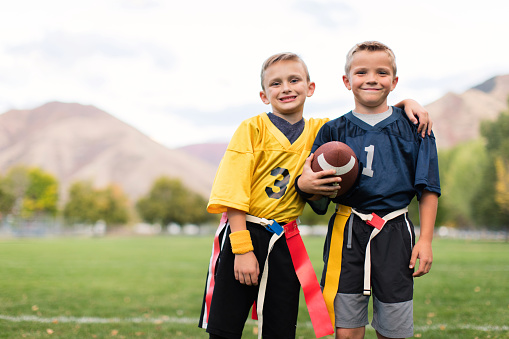 Two young boys dressed in flag football uniforms stand posing for a portrait ready after playing a game. They are smiling and looking at the camera while standing on a football field in Utah, USA.
