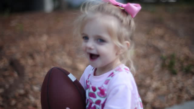 A toddler girl runs around her yard with a football.