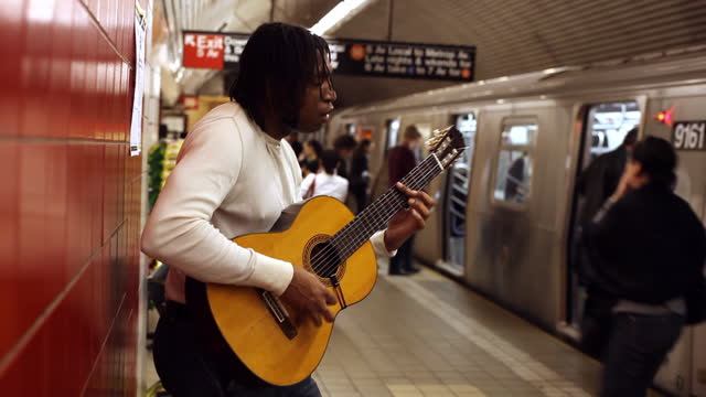 MED profile black man leans against red wall playing guitar in subway station   train enters   passengers board