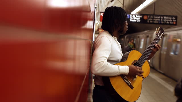 MED profile black man leans against red wall playing guitar in subway station   train leaves   people walk by