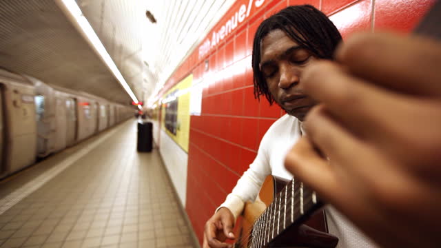 MED black man leaning against red wall plays guitar in subway station   pan to moving train