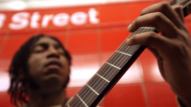 MED black man plays guitar while leaning against red wall in subway station   rack focus