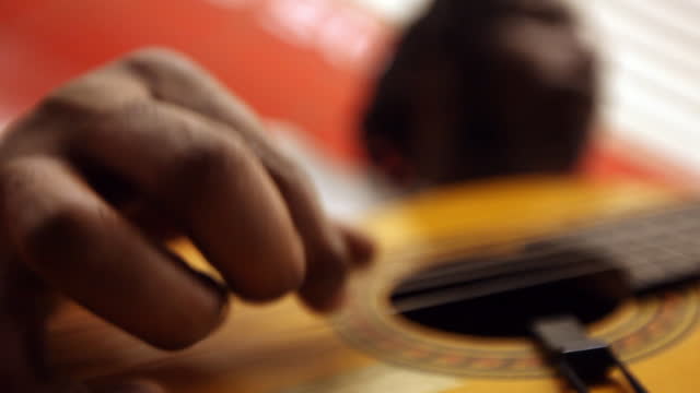 CU fingers of black man playing guitar while leaning against red wall in subway station