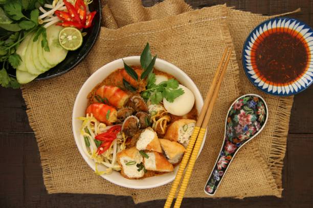 Curry Laksa Curry Laksa, Peranakan spicy noodle soup from Singapore and Malaysia. Egg and rice noodles topped with prawns, cockles, fish balls, tofu, and bean sprouts in fragrant and rich curry broth. Served in a white ceramic bowl; accompanied with chili paste in separate saucer. A pair of wooden chopsticks and a ceramic soup spoon are placed next the bowl. A folded sackcloth  is used as a casual place mat. The meal is set on a rustic wooden dining table. johor photos stock pictures, royalty-free photos & images