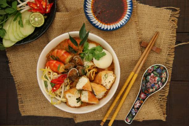 Curry Laksa Curry Laksa, Peranakan spicy noodle soup from Singapore and Malaysia. Egg and rice noodles topped with prawns, cockles, fish balls, tofu, and bean sprouts in fragrant and rich curry broth. Served in a white ceramic bowl; accompanied with chili paste in separate saucer. A pair of wooden chopsticks and a ceramic soup spoon are placed next the bowl. A folded sackcloth  is used as a casual place mat. The meal is set on a rustic wooden dining table. johor photos stock pictures, royalty-free photos & images