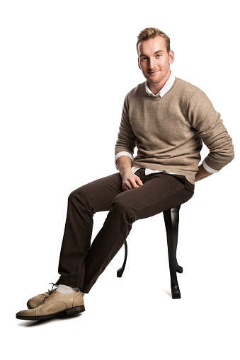 Handsome blonde man wearing a brown pullover and white shirt, sitting down smiling against a white background.