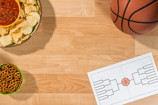 An overhead view of a party for a college basketball tournament with an empty 16 team bracket. There is a bowl of small pretzels, chips and salsa along with a basketball sitting on what looks like a maple wood basketball court. This bracket is for a 16 team single elimination tournament, winner take all,  which typically happens in March every year in the US between college basketball teams and many people gamble on the series.