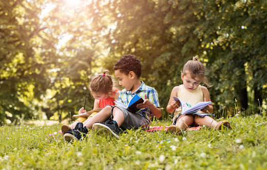 Three small children sitting in grass at the park with their notebooks. Focus is on girl on the right.