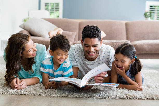 genitori e bambini sdraiati sul tappeto e libro di lettura in soggiorno - domestic life young family family child foto e immagini stock