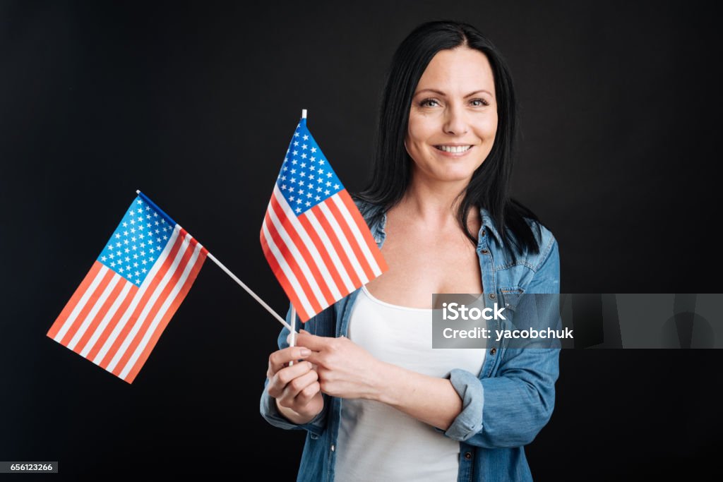 Positive delighted female keeping flags Preparation for celebration. Attractive black-haired woman wearing jeans shirt and white T-shirt keeping smile on her face and posing with two flags in her hands Adult Stock Photo