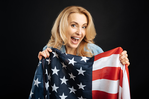 Be proud. Attractive delighted female keeping her mouth opened holding big flag in front of herself posing on black background