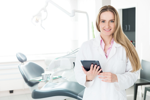 Horizontal color image of female dentist holding digital tablet at modern dental clinic. Young woman wearing white lab coat. Looking at camera and smiling.