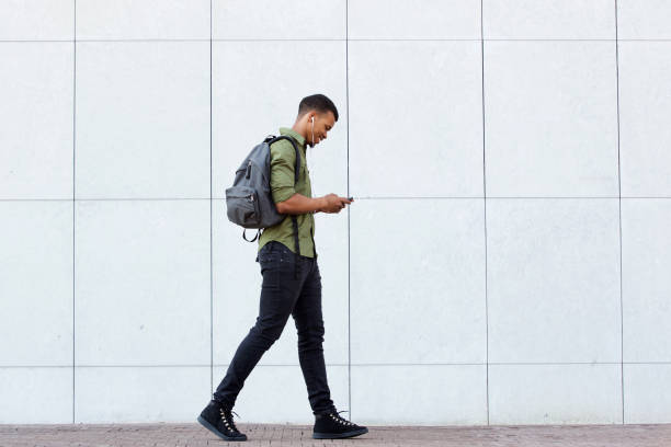 smiling man walking with backpack smart phone and headphones Full length side portrait of smiling man walking with backpack smart phone and headphones men hairstyle afro cool stock pictures, royalty-free photos & images