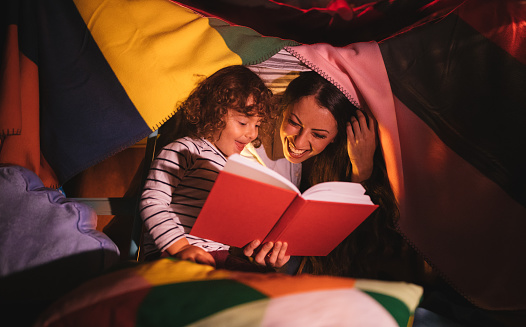 Young mother reading a funny bedtime story to her young boy under blanket fort
