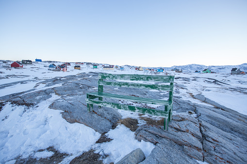 Faded old green wooden bench on rocky outcrop on edge of Greenlandic village. Snowy landscape and colourful houses in background