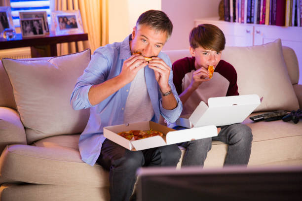 pai e filho comendo pizza e assistindo tv na sala - family sofa night indoors - fotografias e filmes do acervo