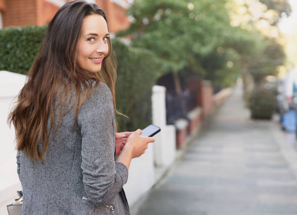 portrait d’une femme d’affaires souriante avec un téléphone portable regardant en arrière sur le trottoir - life back lit people happiness photos et images de collection