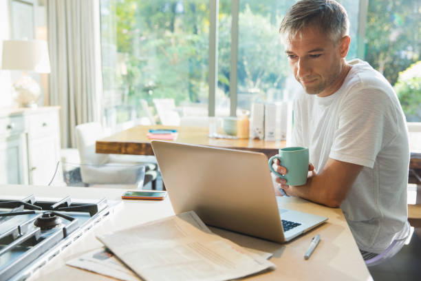 hombre bebiendo café y trabajando en una computadora portátil en la cocina - trabajando en casa fotografías e imágenes de stock