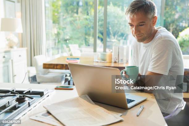 Man Drinking Coffee And Working At Laptop In Kitchen Stock Photo - Download Image Now