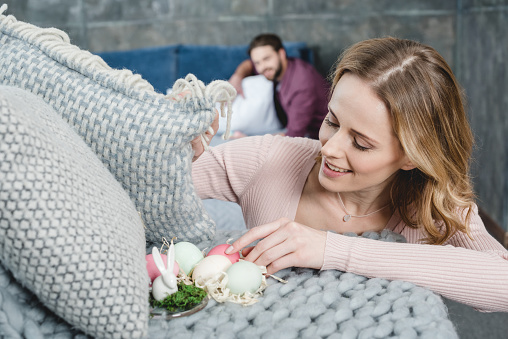 Smiling young woman putting painted easter eggs in decorative nest with shavings