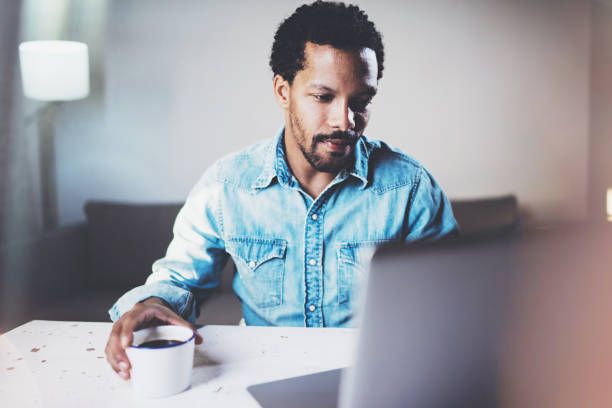 istock Closeup view of pensive bearded African man using laptop at home while drinking cup black coffee on the wooden table.Concept of young people work mobile devices.Blurred background. 656324554
