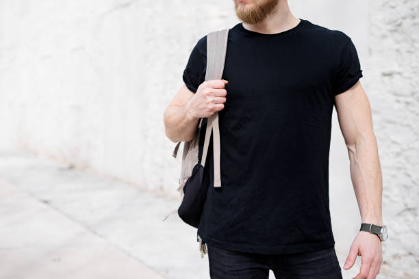Young muscular bearded man wearing black tshirt and backpack posing outside. Empty white concrete wall on the background. Hotizontal mockup, front view