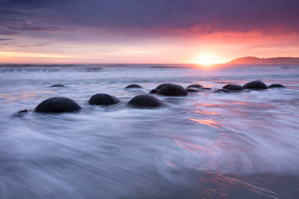 New Zealand. Just after high tide at sunrise, waves break around the Moeraki Boulders. These are large spherical boulders lying on the beach on the Otago coast about 75 km north of Dunedin. Each boulder can be up to 2 meters across.