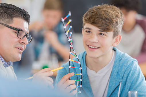 Cheerful Hispanic male student leans about genetics in chemistry class. A teacher is holding a DNA helix model.