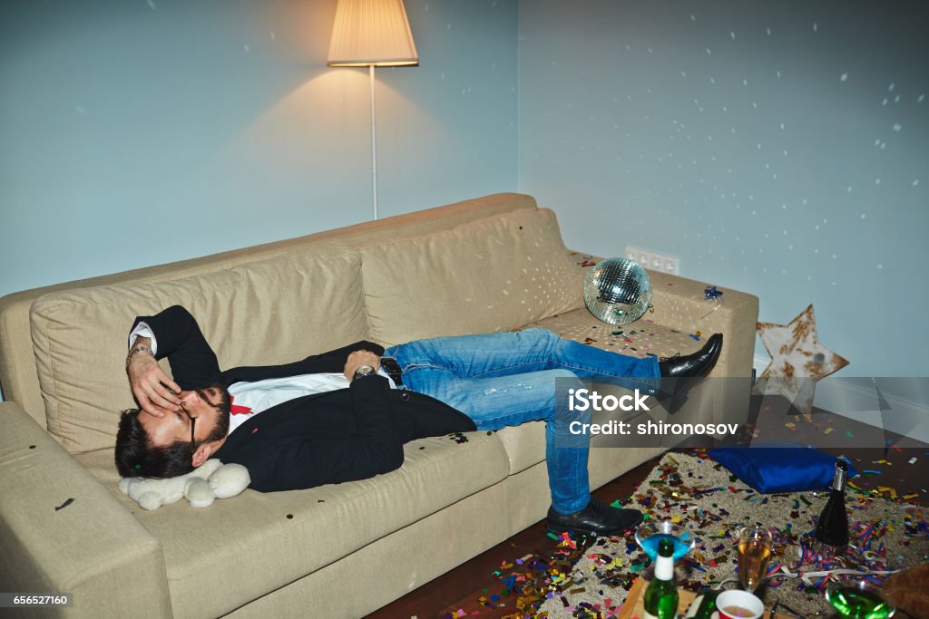 Asian man having hangover after party Asian man in jeans and jacket having nap in messy living room after wild New Year party Flooring Stock Photo