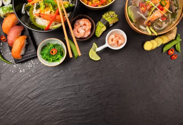 Asian food table with various kind of chinese food, noodles, chicken, prawn soup, rice, sushi and many others. Served on black stone table, top view. copyspace for text