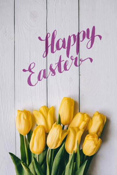 istock Happy Easter hand lettering calligraphy text. Yellow tulips bunch on white wooden planks rustic barn rural table background. Letters, inscription. Beautiful vertical flat lay postcard. 656582886