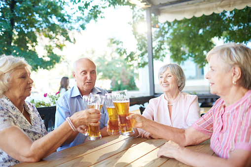 Group of elegant-looking elderly people sitting in outdoor pub and clanging glasses together with joy